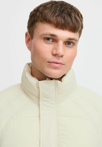 Lys beige pufferjakke med høj krave og lynlås foran, der har en quiltet design og glat tekstur gennem hele jakken.