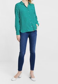 Blouse à pois turquoise avec un design boutonné, manches longues et col, associée à un jean skinny bleu foncé et des talons blancs.