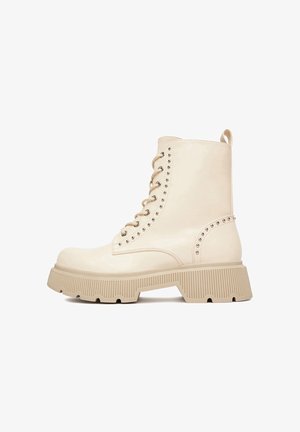 Beige bőr bokacsizma, fűzős előlappal, szegecses díszítésekkel és vastag bordázott talppal a jobb tapadás és stabilitás érdekében.