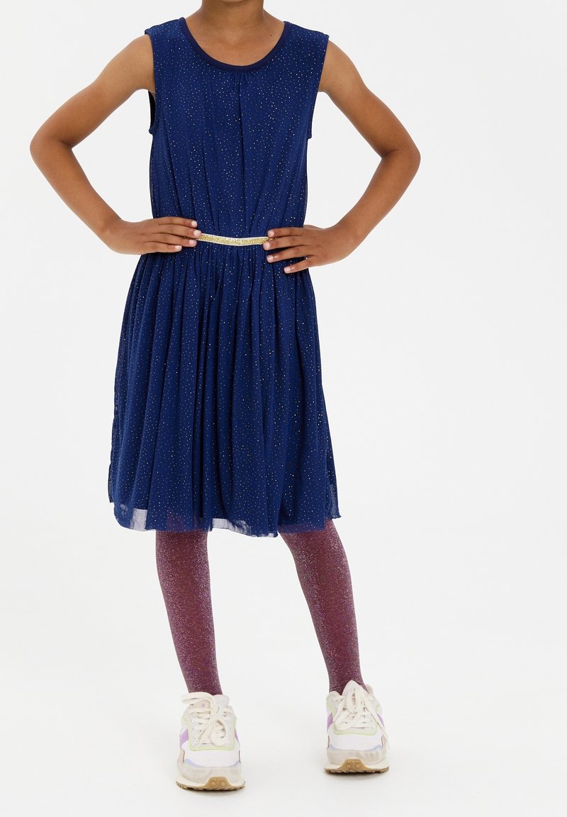 Vestito senza maniche blu navy con punti brillanti, vita arricciata e cintura dorata. Abbinato a leggings lucidi viola e sneakers bianche.