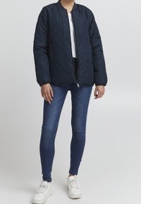 Marineblå quiltet bomberjakke med lynlåslukning, elastiske manchetter og glat tekstur, sat sammen med mørke skinny jeans og hvide sneakers.