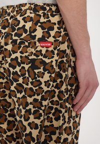 Beige pants with a black and brown leopard print pattern, featuring an elastic waistband and a small red label on the back pocket.