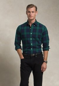 Camisa a cuadros en verde y azul marino con botones, de manga larga y cuello; combinada con pantalones negros y un cinturón. Textura de tela suave.