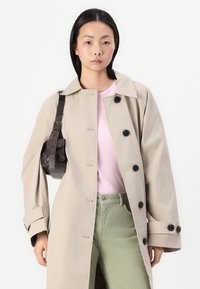 Manteau beige avec des boutons noirs, grand col et manches longues, associé à une chemise rose et un pantalon vert. Sac en bandoulière marron foncé.