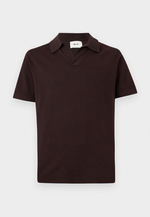 Homme debout portant un polo marron foncé à manches courtes, un jean bleu clair à jambes larges et des sandales noires, avec une main dans la poche sur fond blanc.