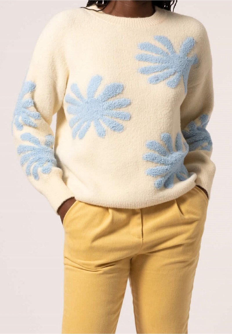Personne portant un pull crème avec des motifs floraux bleus texturés et un pantalon jaune moutarde, mains dans les poches, torse visible.