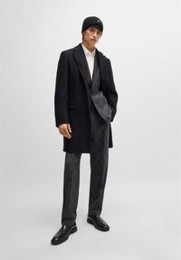 Black overcoat over a gray patterned suit, paired with black leather boots and a black knit beanie. Model stands against a plain background.