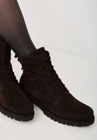 Braune Wildleder-Stiefeletten mit Schnürdesign, strukturiertem Obermaterial, runder Zehenpartie und robustem Profil, mit dunkler Naht und kurzem Schaft.