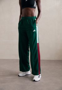 Groene sportbroeken met witte strepen en maroon zijpanelen, met een elastische tailleband en zijzakken. Draagt witte sneakers.
