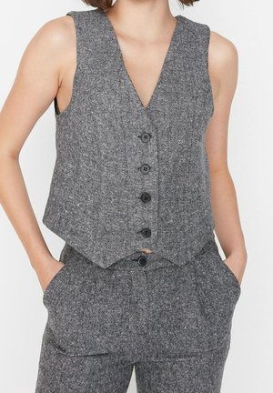 Vrouw die een grijs, mouwloos, geknoopt gilet en bijpassende hoge taille broek draagt, met handen in de zakken, tegen een effen achtergrond.