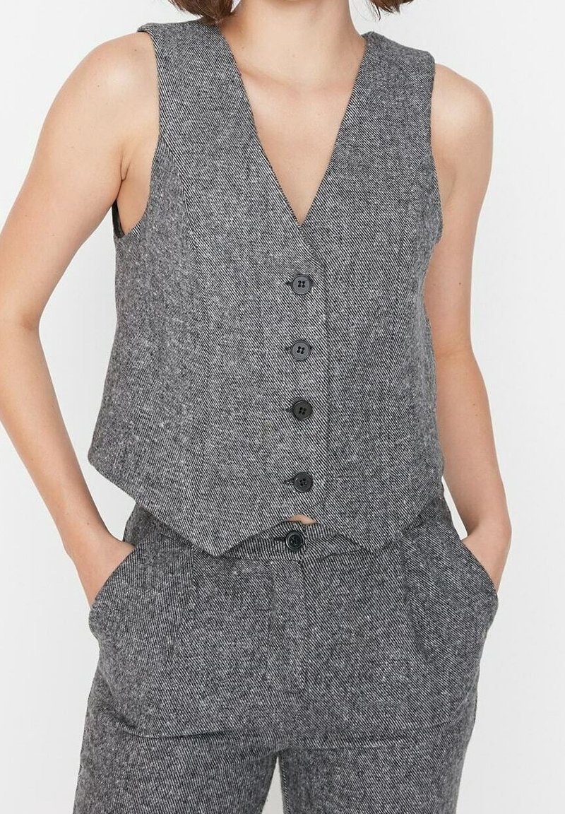 Femme portant un gilet gris sans manches boutonné et un pantalon taille haute assorti, les mains dans les poches, sur un fond uni.