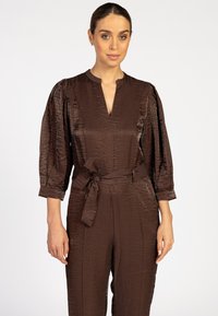 Bruine jumpsuit met textuurstof, opgeblazen mouwen, V-halslijn en een strikriem in de taille. Gladde, glanzende afwerking met geplooide details.