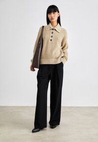 Femme debout portant un pull en maille beige avec un grand col et des boutons, un pantalon noir à jambes larges, des chaussures noires, et portant un sac cabas marron foncé.