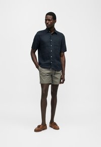 Άνδρας που φοράει σκούρο κοντομάνικο πουκάμισο με κουμπιά, ανοιχτό γκρι σορτς και καφέ δερμάτινα boat shoes, στέκεται με τα χέρια στις τσέπες.