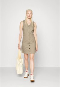 Vestido caqui sem mangas com decote em V, botões frontais e silhueta reta. Acompanhado de uma mala tote bege e ténis brancos.