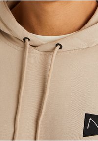 Felpa beige con cappuccio e lacci, occhielli neri e una piccola patch nera con un design astratto bianco sul petto.