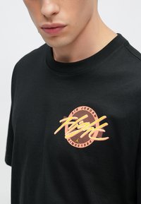 Fekete pamut póló, amelyen egy rózsaszín és sárga kör alakú grafika látható az "AIR JORDAN" és "FLIGHT SINCE 1984" felirattal. Klasszikus kerek nyakú kialakítás.