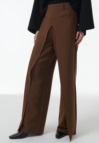Pantalons évasés marron avec un pli marqué, dotés d'une taille haute et d'une poche zippée sur le côté. Texture lisse et design ajusté.