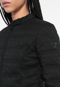 Must tikk-kangaga jope kõrge kraega, millel on peen logo muster, tõmblukk ja kolmnurkne brändi märk varrukal.