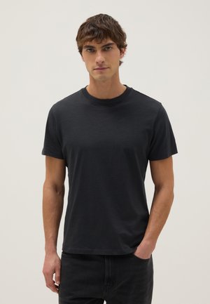 Giovane uomo con capelli castani corti che indossa una semplice maglietta nera a maniche corte e pantaloni neri, in piedi su uno sfondo bianco.