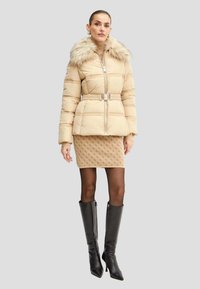 Beige puffjakke med faux fur-krave, bælte detaljer og skrå stikninger. Båret over en struktureret beige nederdel og parret med knæhøje sorte støvler.