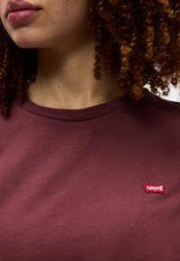 Κοντινό πλάνο ενός ατόμου με σγουρά μαλλιά που φοράει πουκάμισο μαρουν  χρώματος Levi’s με μικρό κόκκινο λογότυπο στο στήθος.