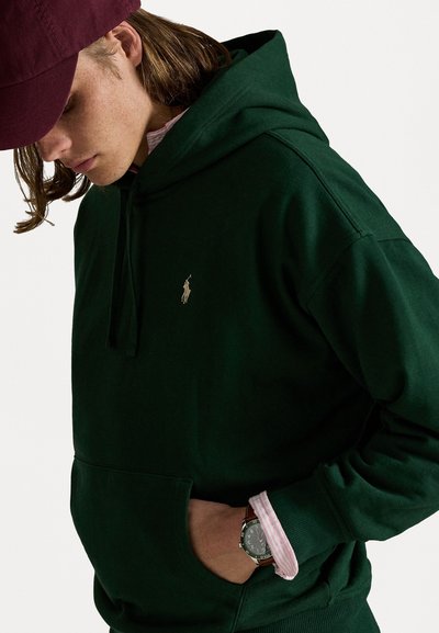 Grøn bomulds hoodie med en frontlomme og hætte med snor, prydet med et lille, gyldent logo. En pink stribet skjorte kan ses under. 