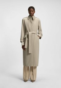 Beiger Trenchcoat mit tailliertem Gürtel, langen Ärmeln und Manschettendetails. Getragen über beigen weit geschnittenen Hosen mit spitzen Schuhen.
