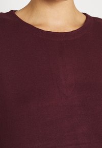 Persona che indossa un maglione girocollo di colore viola scuro, con il colletto e la parte superiore del petto in evidenza su uno sfondo neutro.