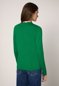 Zielona koszula z długim rękawem i zdobieniem w postaci falbanki przy kołnierzu. Wykonana z gładkiej tkaniny, ma dopasowany fason i jest zestawiona z niebieskimi dżinsami.