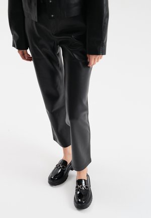 Black leather straight-cut trousers paired with glossy black loafers featuring a silver chain accent. Smooth texture throughout.