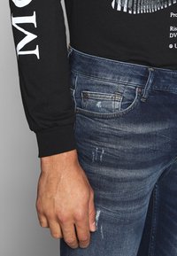 Camisa negra de manga larga con texto blanco; jeans de mezclilla azul con un acabado desgastado, desgarros y detalles de costura visibles. Mano descansando sobre los jeans.