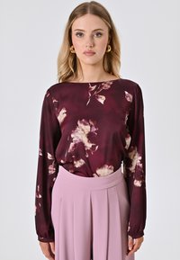 Langärmlige bordeauxfarbene Bluse mit Blumenmuster und Rundhalsausschnitt, kombiniert mit hoch taillierten, plissierten Hosen in zartem Rosaton.