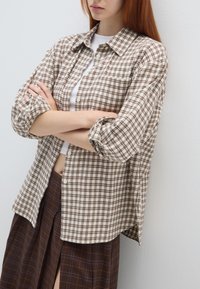 Camicia a quadri marrone e crema con bottoni, maniche lunghe, vestibilità rilassata; indossata sopra un top bianco e una gonna a quadri marroni.