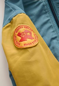 Kleurvolle jas met een blauwe bovenkant, gele onderkant en een rode geborduurde patch op de mouw met een sterdesign.