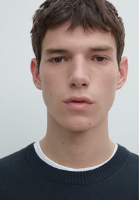 Ciemny dzianinowy sweter z prążkowanym dekoltem, noszony na białej koszulce z okrągłym kołnierzem, charakteryzujący się gładką fakturą i minimalistycznym designem.