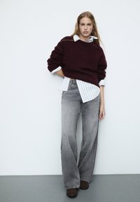 Burgundski oversized pulover složen preko bijele majice s prugama, uparen s svijetlosivim trapericama s trapez krojem i smeškim čizmama do gležnja. Minimalistička pozadina.