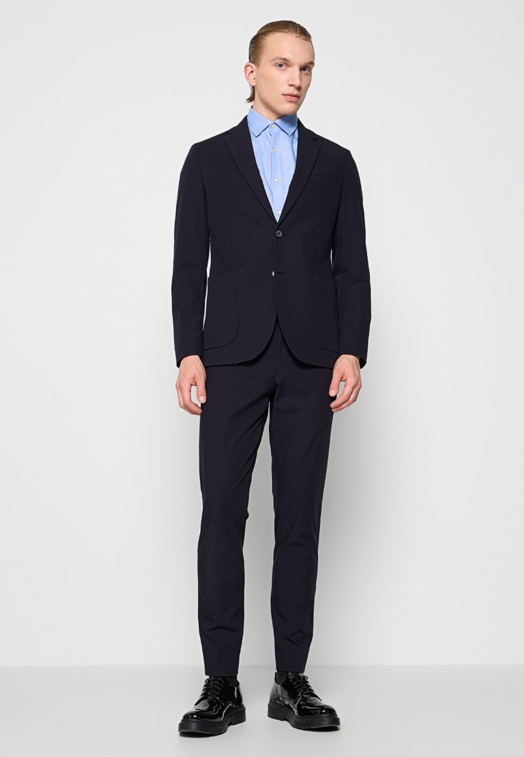 Marineblauer maßgeschneiderter Anzug mit einem Ein-Knopf-Blazer, passenden schlanken Hosen und einem hellblauen Hemd darunter. Glänzende schwarze Schuhe.