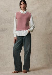 Top sans manches en tricot rose superposé sur une chemise blanche à boutons, associé à un jean baggy gris foncé et des chaussures plates marron. Design simple et texturé.