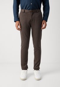 Brown patterned trousers with a slim fit, featuring a waistband and a button closure. Worn with white sneakers for contrast.