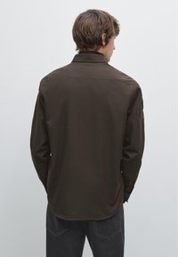 Chemise marron à manches longues avec une coupe décontractée, un design minimaliste et un col classique, présentant une texture lisse et un ourlet droit à l'arrière.