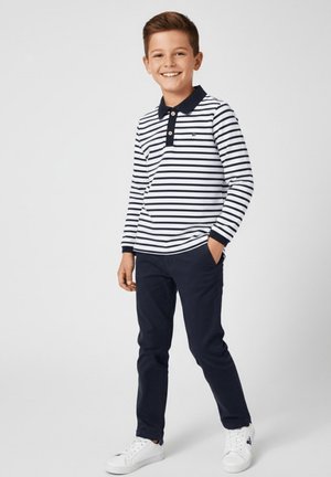 Chłopiec uśmiechający się, ubrany w białą koszulę z długim rękawem w granatowe paski, granatowe spodnie, białe trampki, stojący z jedną ręką w kieszeni na jednolitym tle.