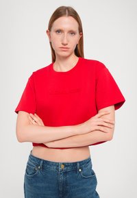 Tricou scurt roșu, confecționat din bumbac, cu mâneci scurte, purtând logo-ul reliefat "MICHAEL KORS" pe partea din față. Asortat cu blugi albaștri.
