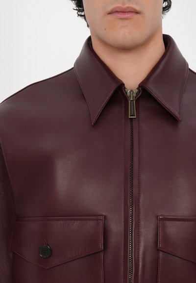 Bordeauxfarbene Lederjacke mit spitzem Kragen, vorderem Reißverschluss und zwei großen, flachen Taschen mit Knopfakzenten.