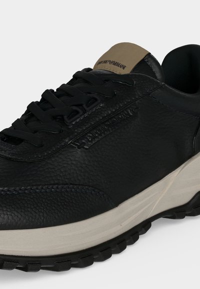 Μαύρο sneaker από υφή δέρματος με μαύρα κορδόνια, μπεζ επενδυμένη γλώσσα και λευκή σόλα με μαύρα σχέδια πέλματος.