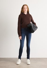 Brown knit sweater with hood, ribbed texture; dark blue skinny jeans; holding a black leather bag; white sneakers. Neutral background.