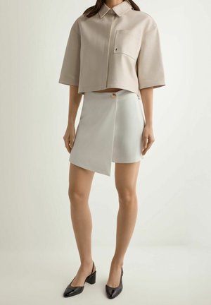 Kobieta w beżowej krótkiej koszuli z kieszonką, białej asymetrycznej mini spódnicy z guzikiem oraz czarnych sandałach na obcasie z paskiem z tyłu.