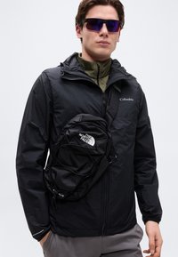 Fiatal férfi tükröződő napszemüveget visel, fekete Columbia dzsekit, alatta olívazöld cipzáras felsőt, szürke nadrágot és fekete The North Face oldaltáskát.