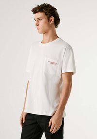 Camiseta de algodón blanca con mangas cortas, cuello redondo y bolsillo en el lado izquierdo con texto rojo: "Nacido para ser una estrella del rock, obligado a trabajar."