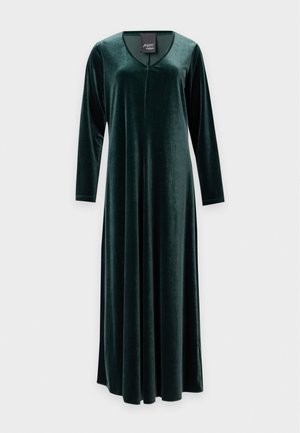 Femme aux longs cheveux blonds portant une robe midi en velours vert foncé à manches longues et des talons noirs, debout devant un fond blanc.